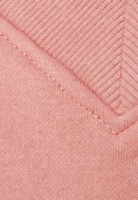 Jasnoróżowa tkanina swetra o texture dzianiny i wzorze w zygzak w górnej części. Posiada gładkie wykończenie i jednolity splot.