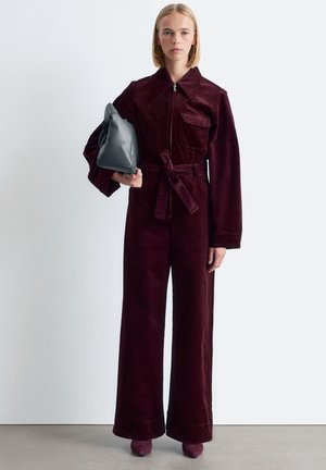 BurgunderfarbenerCord-Jumpsuit mit Frontreißverschluss, großem Kragen und tailliertem Schnitt. Das Modell trägt eine graue Tasche, die mit passendem Schuhwerk kombiniert ist.