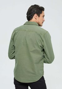Chemise longue en coton verte, à manches longues, avec un col pointu et un ourlet arrondi. Présente deux coutures d'épaule au dos et une texture lisse.