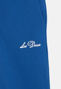 Blaue Stoff-Jogger mit glatter Textur, Seitentaschen und einem weißen gestickten Logo "Les Deux" auf der Vorderseite.