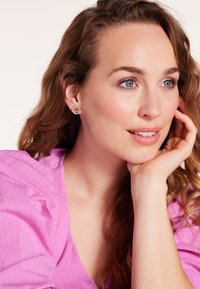 Goudkleurige oorbellen met een bloemmotief, in een ronde vorm, hebben een centrale oranje steen, omringd door kleine heldere stenen. Zijde roze blouse.