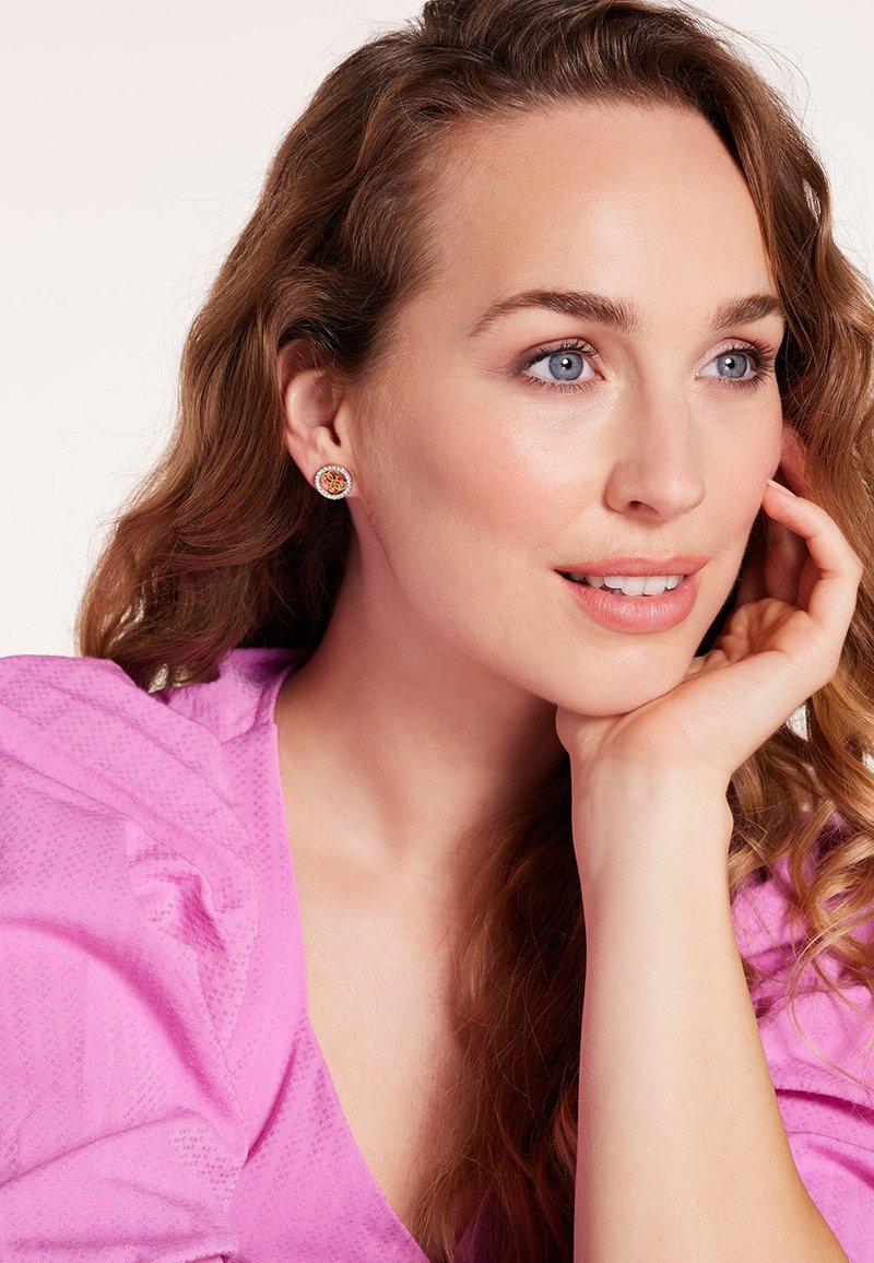 Goudkleurige oorbellen met een bloemmotief, in een ronde vorm, hebben een centrale oranje steen, omringd door kleine heldere stenen. Zijde roze blouse.