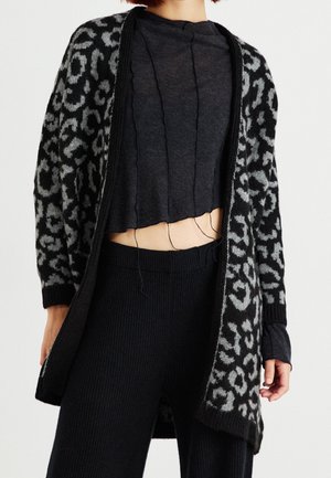 Femme portant un cardigan imprimé léopard noir et gris, un top court gris foncé avec des fils lâches, et un pantalon noir côtelé.