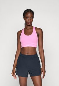 Femme athlétique se tient de face, portant un soutien-gorge de sport rose et un short noir, devant un fond clair et uni.