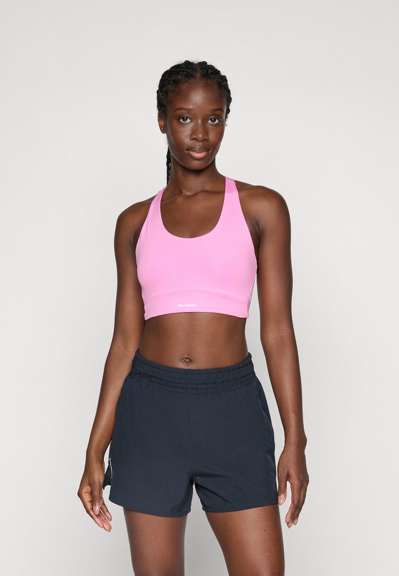 Femme athlétique se tient de face, portant un soutien-gorge de sport rose et un short noir, devant un fond clair et uni.