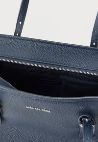 Merensininen nahkalaukku, jossa on kupruileva pinta, hopeinen vetoketju ja logoyksityiskohta. Yläosa on avoin ja sisältä löytyy tasku.