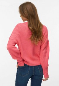 Žena s dugom smeđom kosom u ružičastom pletenom džemperu i plavim trapericama, okrenuta leđima s rukama u stražnjim džepovima.