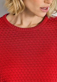 Eine Frau mit blonden Haaren, die einen strukturierten roten Strickpullover trägt und leicht nach unten und zur Seite schaut.