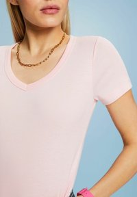Světle růžové přiléhavé tričko s výstřihem do V a krátkými rukávy. Výrazně hladká textura; nošeno s řetízkovým náhrdelníkem a růžovým náramkovým hodinami.