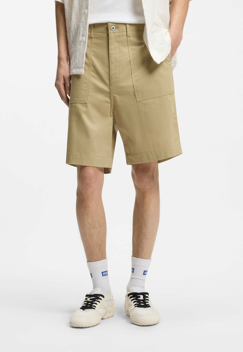 Person trägt beige kniehohe Shorts mit großen Fronttaschen, weiße Socken mit blauem Logo und weiße Turnschuhe mit schwarzen Schnürsenkeln.