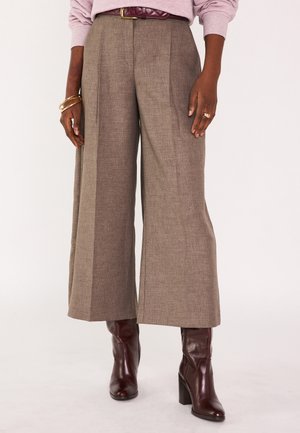 Femme portant un pantalon taupe à jambes larges avec une ceinture bordeaux et des bottes à talons hauts en cuir assorties, accessoirisée avec des bracelets et une bague.