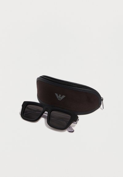 Μαύρα ορθογώνια γυαλιά ηλίου με σκούρα φακούς, τοποθετημένα μπροστά από καφέ θήκη με φερμουάρ και ασημένιο λογότυπο.