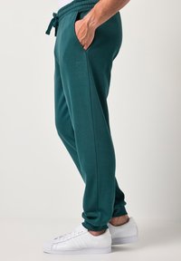 Tyrkysové tepláky se stahováním v pase, bočními kapsami a žebrovanými manžetami, doplněné bílými teniskami. Na kalhotách je nenápadné logo.