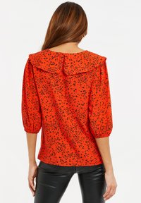 Rode blouse met zwarte pantervlekken, uitlopende kraag, pofmouwen en knoopsluiting op de rug. Glad stof en losse pasvorm.