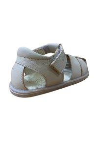 Sandal de cuero beige con parte trasera abierta, tira de velcro y recortes laterales. Presenta una suela lisa y ligero acolchado en el interior.