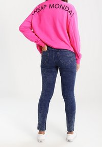 Sudadera rosa de gran tamaño con "CHEAP MONDAY" impreso en la parte de atrás, combinada con vaqueros ajustados azul oscuro y zapatillas blancas.