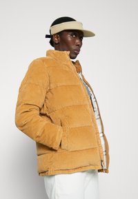 Chaqueta de plumas de corduroy en mostaza con secciones acolchadas horizontales, bolsillos laterales y un cuello alzado. Usada con una visera beige.