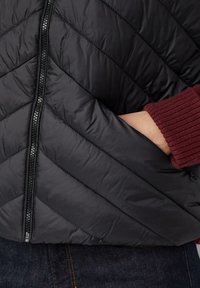 Hand in der Tasche einer schwarzen, gesteppte Jacke mit Frontreißverschluss, die über dunkler Jeans getragen wird und mit einem roten, gerippten Strickpulloverärmel kombiniert ist.