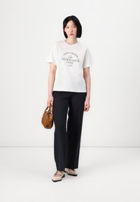 T-shirt blanc en coton avec impression noire, pantalon ajusté bleu marine, ballerines beiges avec brides noires et un petit sac à main marron. Design simple et classique.
