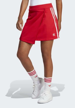 Vrouw met een rode Adidas rok met witte strepen, witte Adidas sokken met rode strepen en witte sneakers tegen een effen achtergrond.