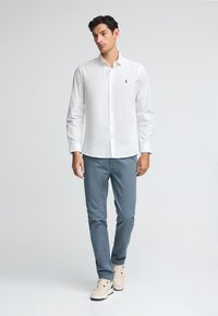 Vit skjorta med knappar i tyg, med en liten logotyp på bröstet, kombinerad med ljusblå byxor och beige sneakers.