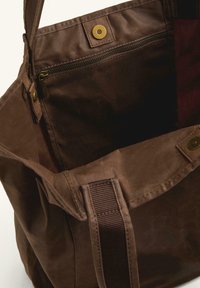 Brown canvas tote bag with a top snap closure, featuring an interior zip pocket and sturdy fabric straps. Smooth texture, spacious design.