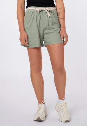 Persoon die groene shorts met hoge taille draagt met een beige stoffen riem, zwart topje, witte sokken en witte sneakers, staand tegen een effen achtergrond.