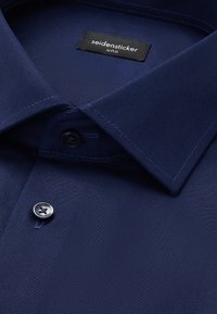 Granatowa koszula z klasycznym kołnierzykiem, pojedynczym zapięciem na guzik oraz tekstylną metką z napisem "seidensticker uno." Gładka faktura tkaniny.