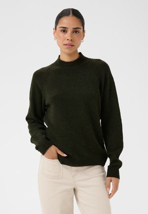 Femme aux cheveux foncés portant un pull vert foncé et un pantalon beige clair, debout, une main dans la poche, sur un fond uni.