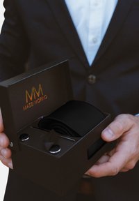 Must mustriba mustravööt, mis on mattmustas karbis kuldse logoga. Karp on varustatud kahe ümmarguse kinnitusega ja musta paelaga tõmbeelemendiga.