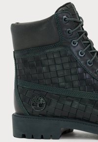 Black leather boot with woven texture, rubber sole, green stitching, and round metal eyelets. Features logo near the base.