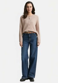 Hellrosa Strickjacke mit Knopfleiste und langen Ärmeln, kombiniert mit dunkelblauen Jeans mit weitem Bein und einem braunen Gürtel, getragen mit schwarzen Schuhen.