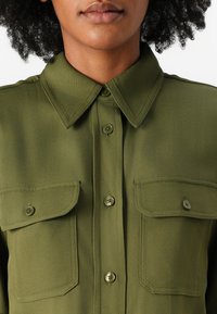 Camicia verde oliva, con bottoni, realizzata in tessuto liscio con due tasche sul petto. Presenta un colletto a punta e bottoni coordinati.