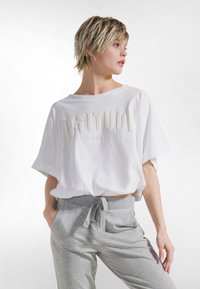 Camiseta blanca recortada con texto "ACTITUD" en letras suaves y con textura. Combinada con pantalones deportivos gris claro con cintura ajustable con cordón.