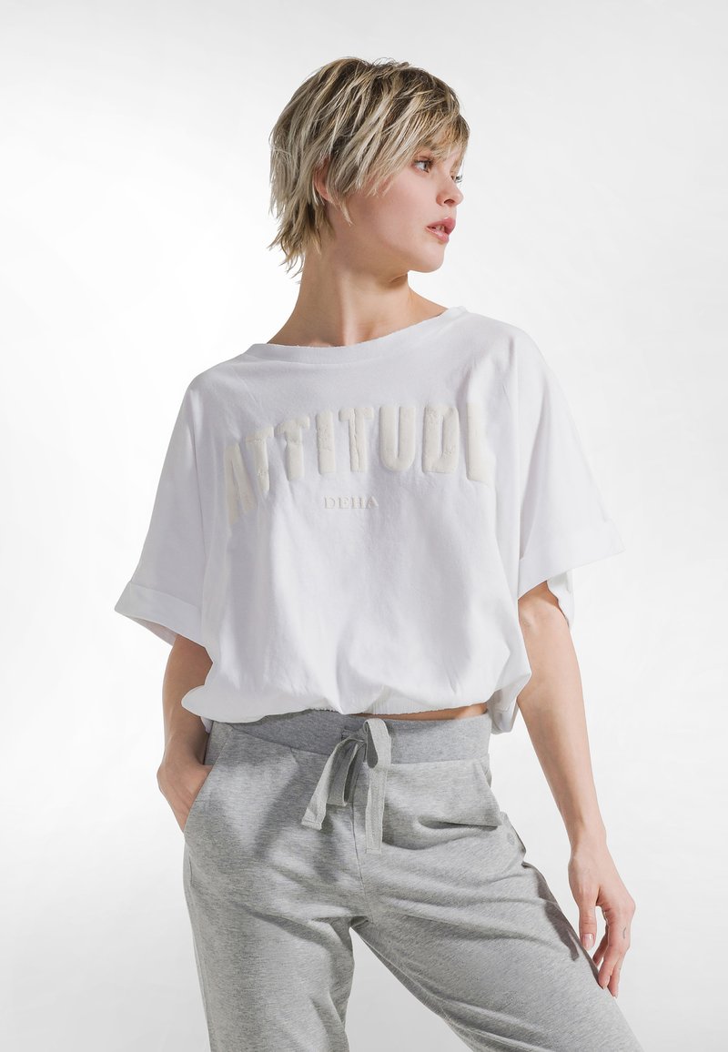 Camiseta blanca recortada con texto "ACTITUD" en letras suaves y con textura. Combinada con pantalones deportivos gris claro con cintura ajustable con cordón.