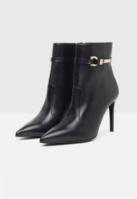 Black leather ankle boots with pointed toes, stiletto heels, and a gold accent band. Smooth texture and sleek, modern design.