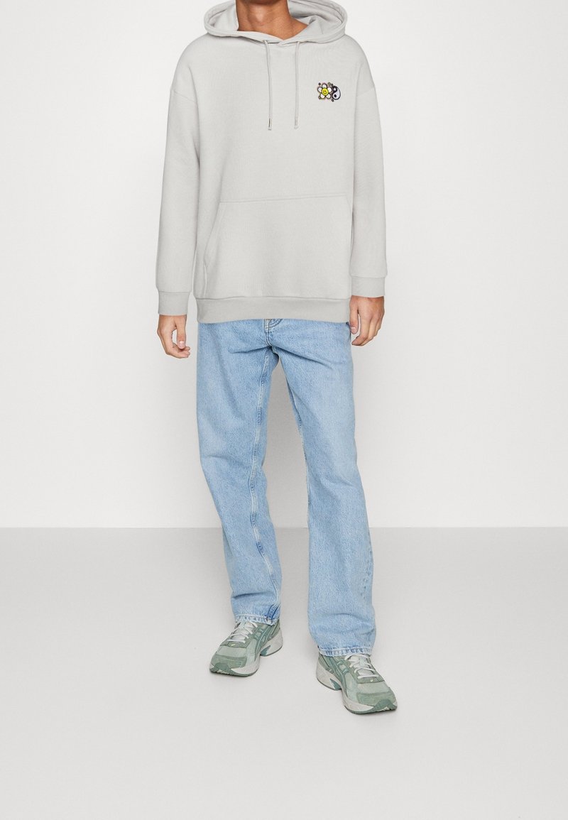 Personne portant un sweat à capuche gris clair avec un petit logo fleur souriante sur la poitrine, un jean bleu clair et des baskets vert-gris debout.
