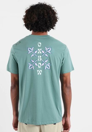 T-shirt en coton vert avec un grand motif graphique blanc et lavande au dos, comprenant le mot "Oxbow" et des motifs de feuilles.
