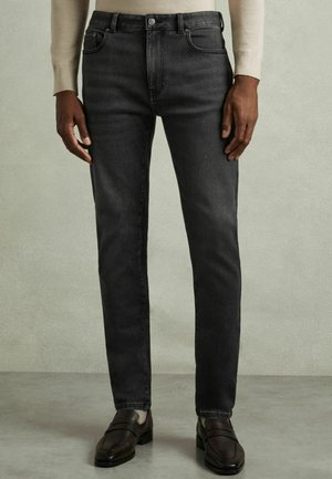 Mand iført slimfit sorte jeans, beige sweater og mørkebrune læder loafers står på en grå gulv mod en lys væg.