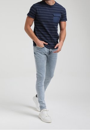 Mand iført en marineblå stribet T-shirt med brystlomme, lyseblå skinny jeans og hvide sneakers, går med hænderne tæt på lommerne.