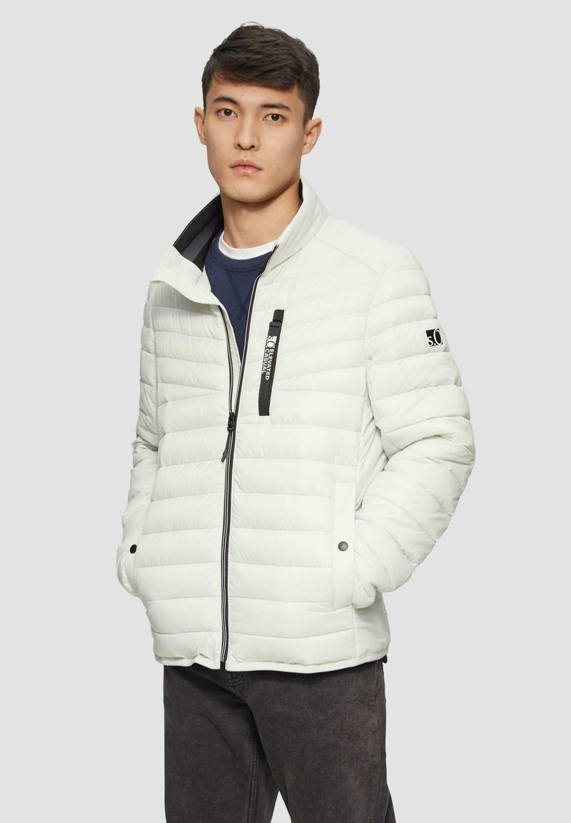 Weiße gesteppte Jacke mit hohem Kragen, Reißverschluss vorne, schwarzen Akzenten auf der Brust und seitlichen Taschen. Getragen über einem marinefarbenen Hemd und dunklen Hosen.
