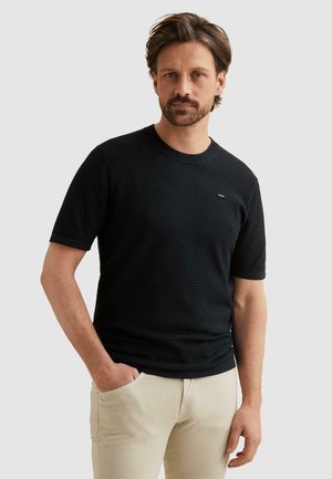 Mann mit Bart, der ein schwarzes, strukturiertes Kurzarmhemd und hellbeige Hosen trägt, steht mit einer Hand in der Tasche vor schlichtem Hintergrund.