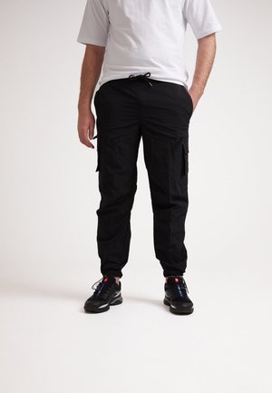 Black cargo pants with multiple side pockets, textured fabric, elastic waistband with drawstring, and tapered cuffs, paired with black sneakers.