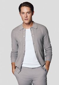 Jasnoszary kardigan uszyty z miękkiego materiału, z zapięciem na guziki, kołnierzykiem oraz długimi rękawami, noszony na białej koszuli i dopasowanych szarych spodniach.