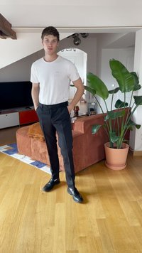 White t-shirt, black tailored pants, and black shoes. Background features a brown corduroy sofa and large potted green plant. Wood flooring.