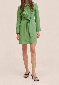 Abito camicia verde con vita regolabile, maniche lunghe e chiusura con bottoni davanti. Tessuto morbido con vestibilità comoda, abbinato a sandali neri con tacco.