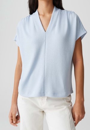 Femme portant un haut texturé bleu clair à manches courtes et col en V ainsi qu'un pantalon blanc taille haute, debout devant un fond neutre.
