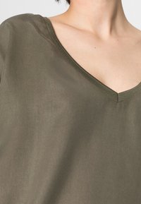 Top verde oliva a scollo a V realizzato in tessuto liscio, con una consistenza morbida e un design pulito e minimalista senza decorazioni.