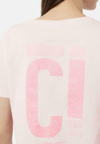 Helles rosa T-Shirt mit einem großen, strukturierten pinken Grafikmotiv auf der Rückseite, das die Buchstaben "CI" und "EST 2010" in einer lässigen Schriftart zeigt.