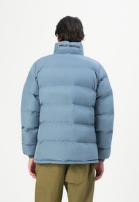 Mężczyzna w jasnoniebieskiej ocieplanej kurtce z napisem "Helly Hansen" na kołnierzu, w zestawieniu z oliwkowymi spodniami, zwrócony plecami w kierunku białego tła.